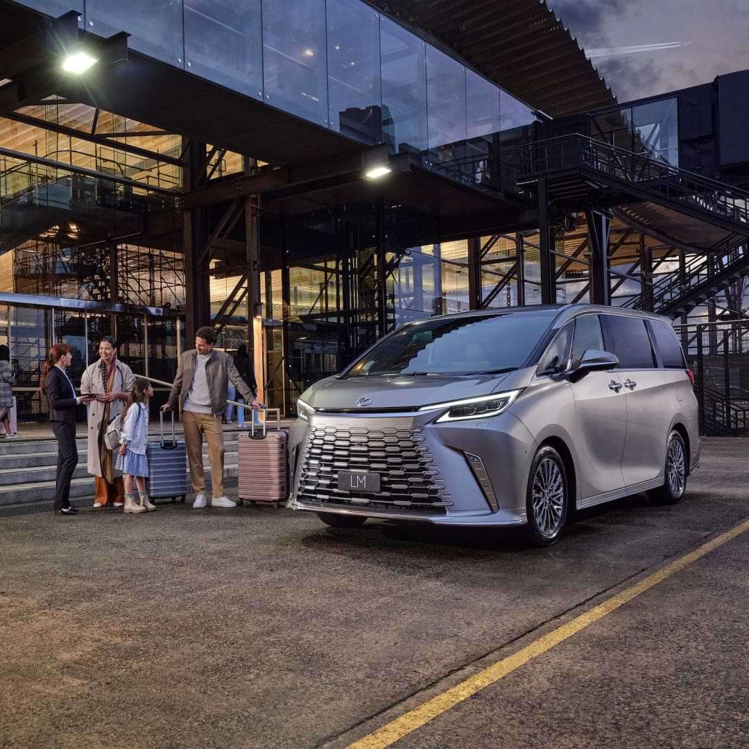 A family loads luggage into a silver Lexus LM at an airport arrivals zone.