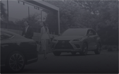 Two people stand between a black sedan and a silver Lexus NX SUV parked outdoors.