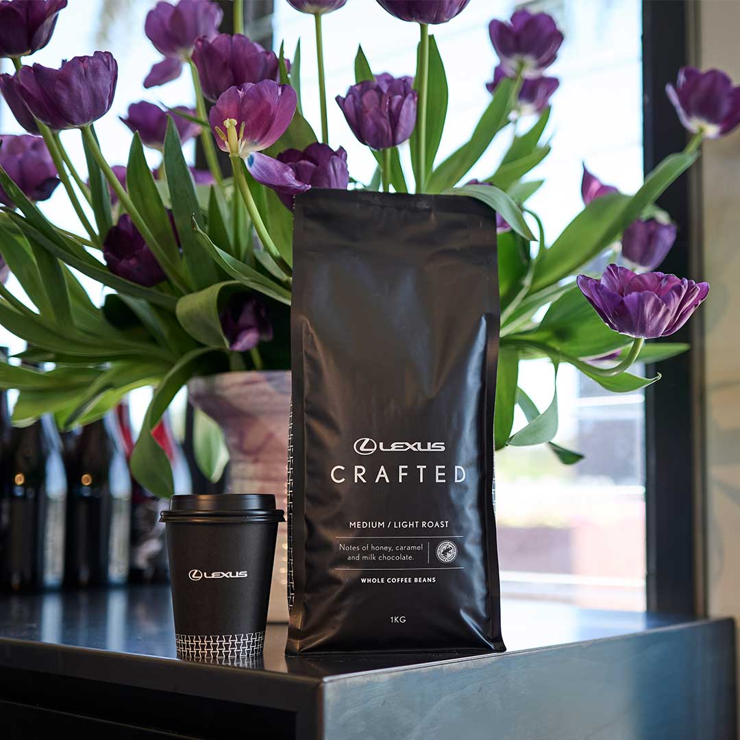 A bag of Lexus Crafted coffee beans and a takeaway coffee cup displayed on a counter beside a vase of purple flowers.