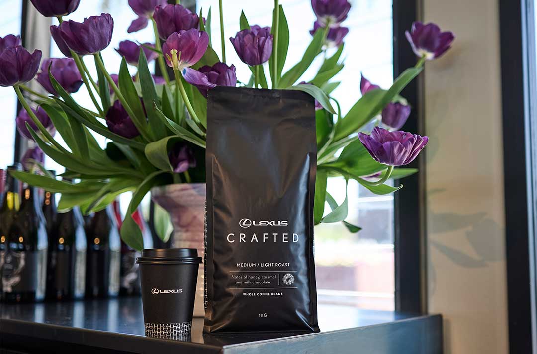 A bag of Lexus Crafted coffee beans beside a takeaway coffee cup and a vase of purple flowers on a counter.