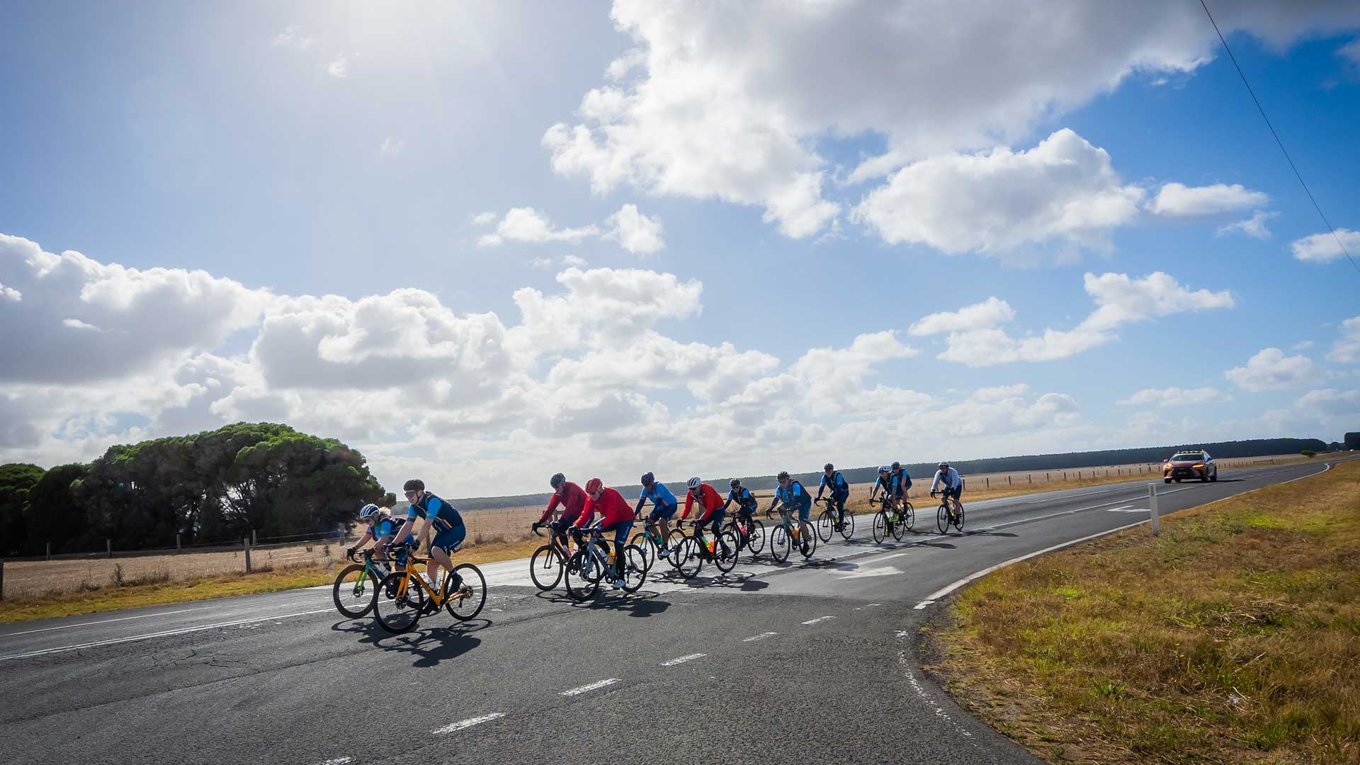 A line of cyclists ride together along an open road through farmland.