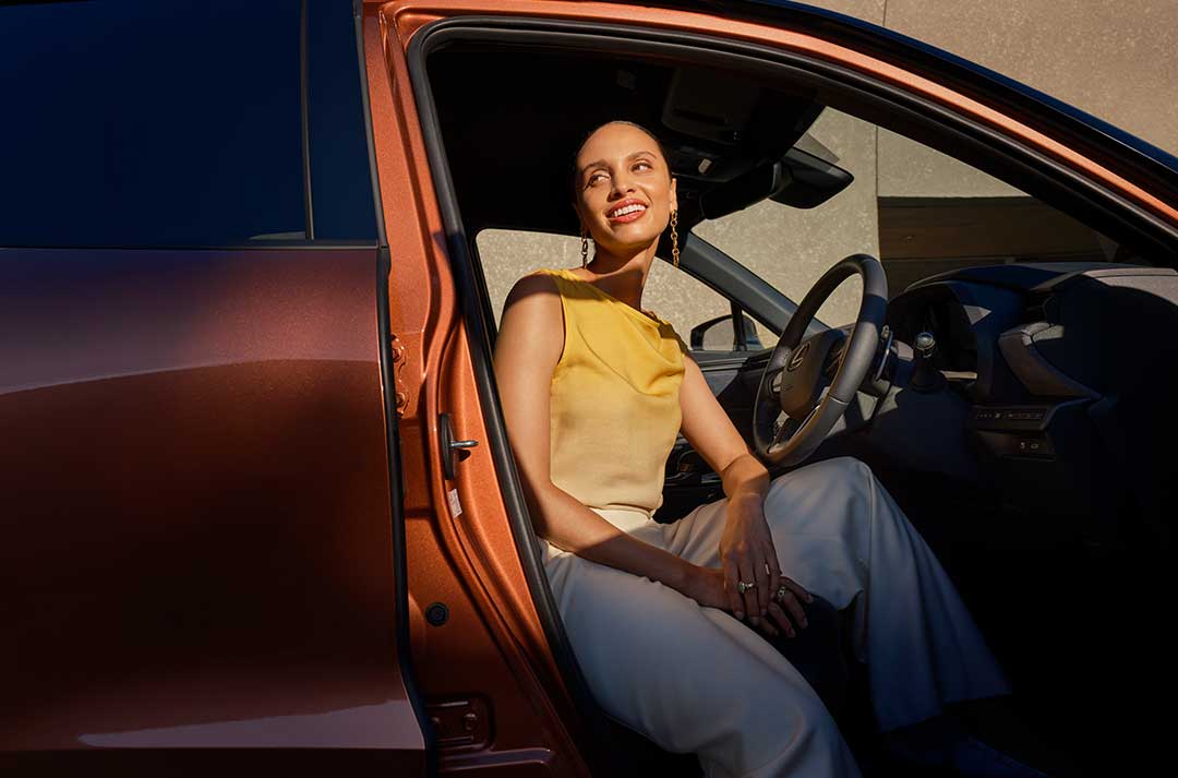Woman sitting in a copper Lexus car's driver's seat with the car door open.