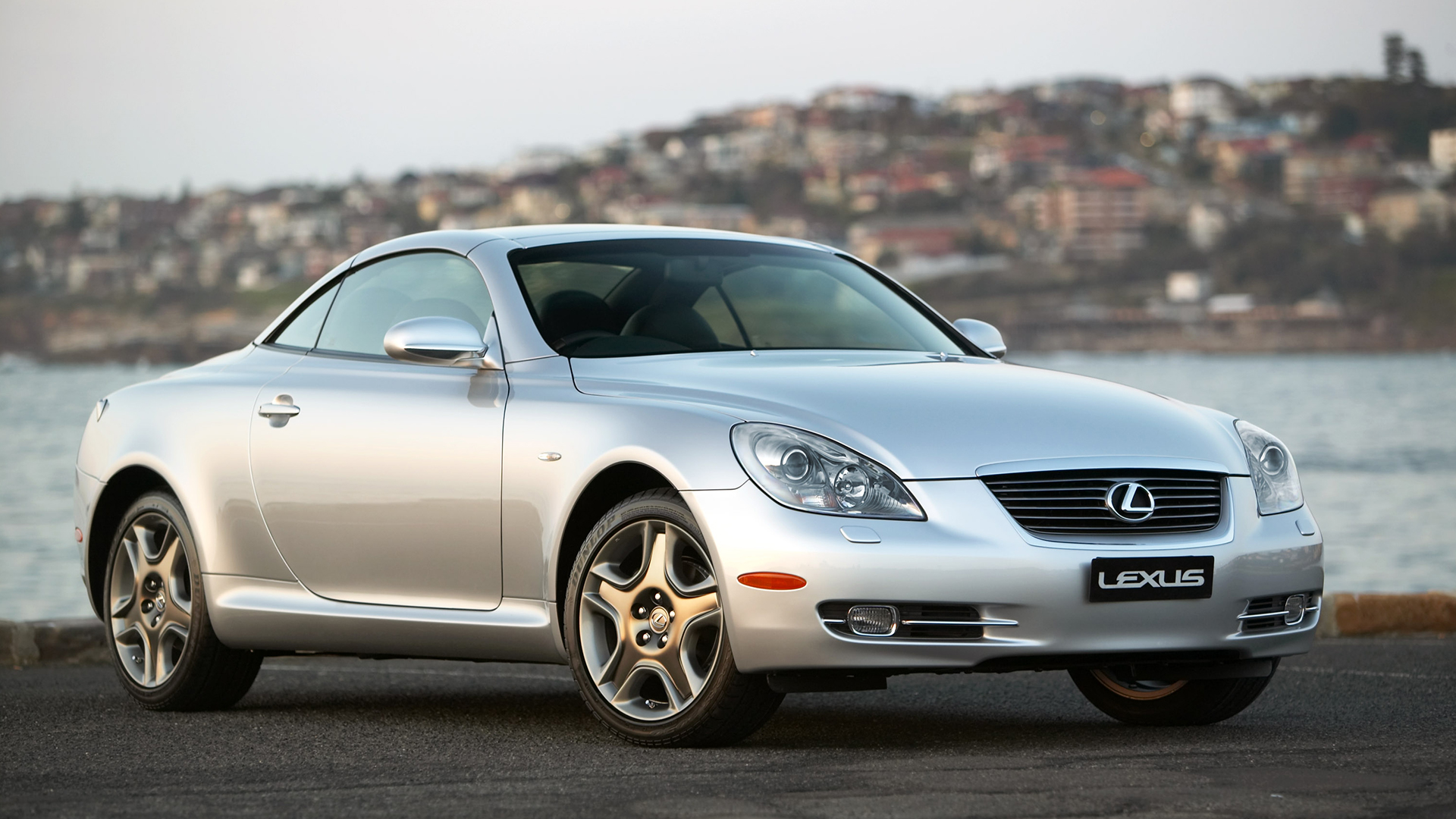 Silver Lexus SC 430 parked on a road with a bay and houses in the background