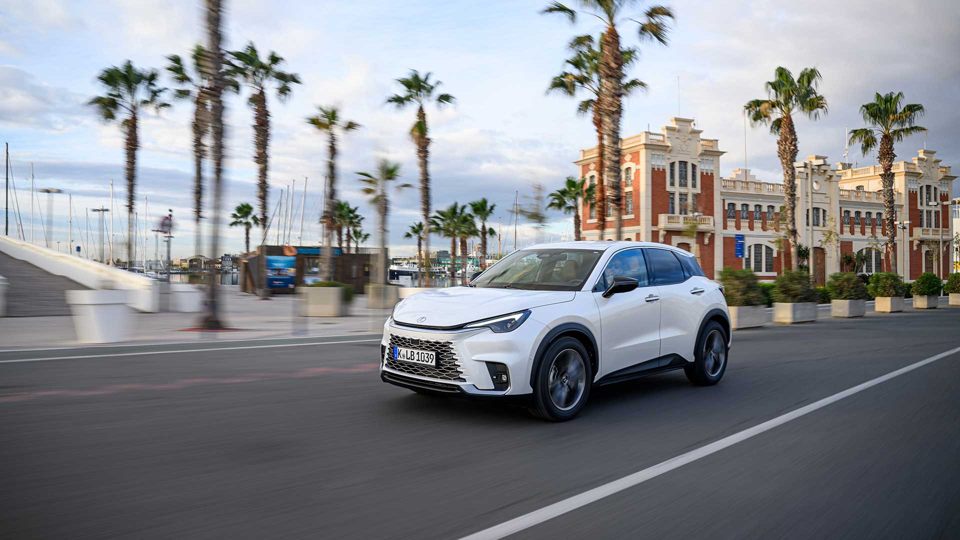 A white Lexus LBX drives on a road with palm trees in the background