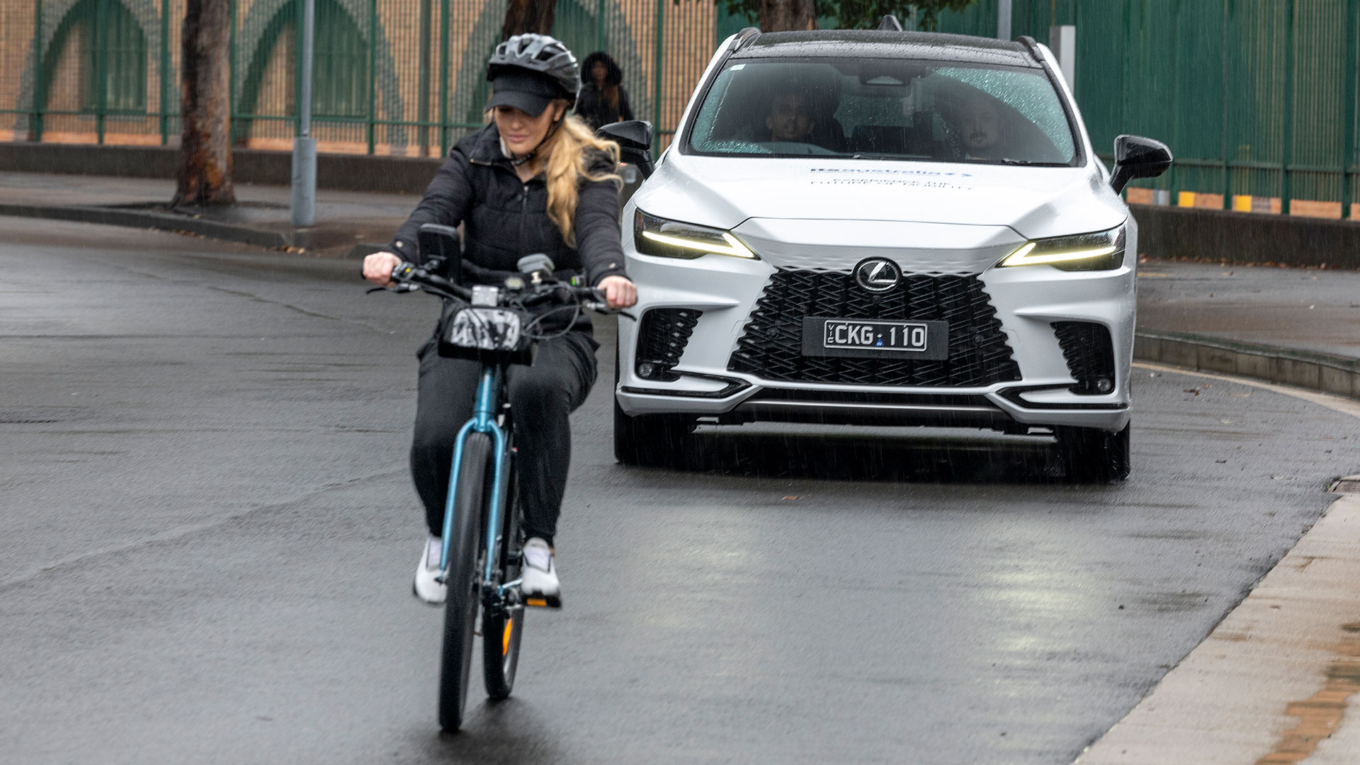 A woman rides a bicycles in front of a white Lexus vehicle
