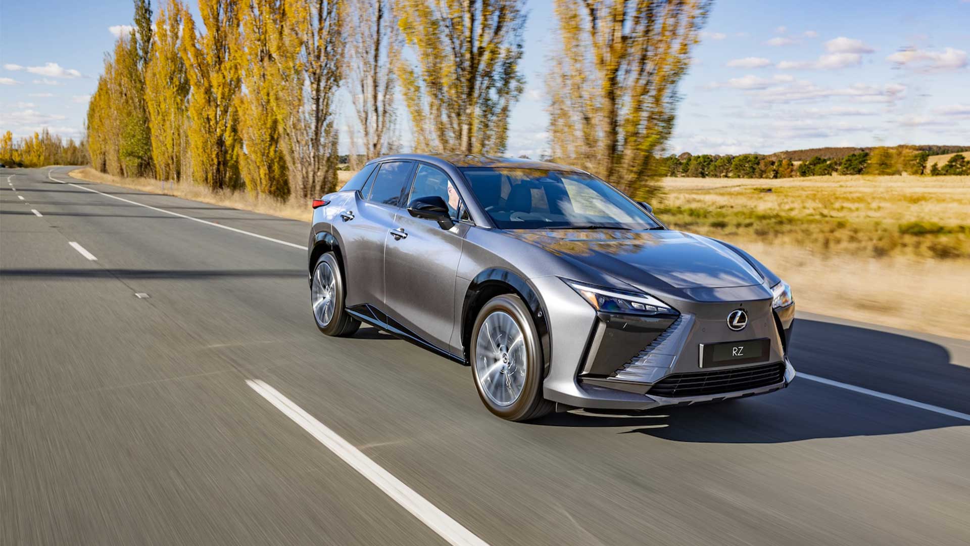 Grey Lexus RZ drives along a landscape with trees and a blue sky in the background