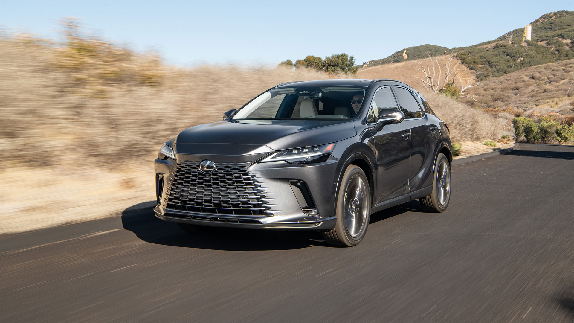 A black Lexus RX 450h+ plug-in hybrid vehicle driving along a road with hills in the background