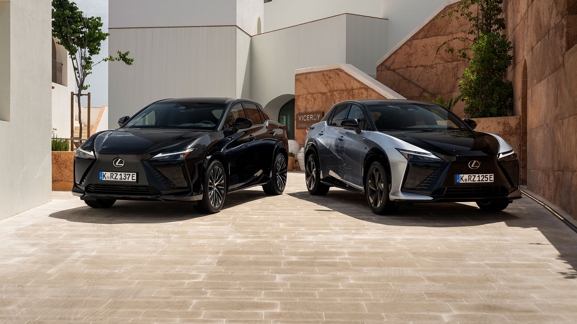 Two Lexus RZ electric SUVs, black and silver, parked side by side in a modern architectural courtyard
