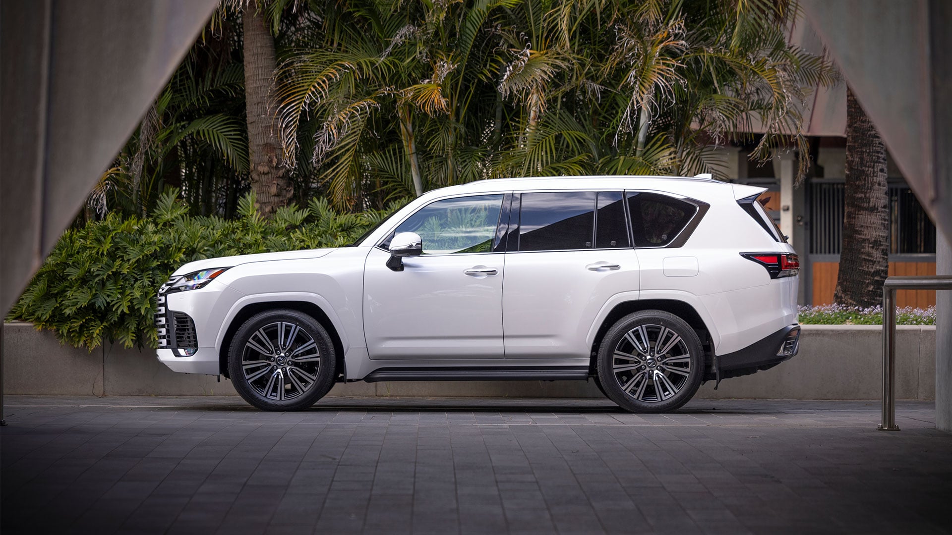 A white 2025 Lexus LX 600 Sports Luxury parked outside a modern building surrounded by trees