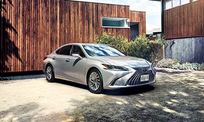 Silver Lexus ES compact crossover parked at an angle in front of a modern, wooden house.
