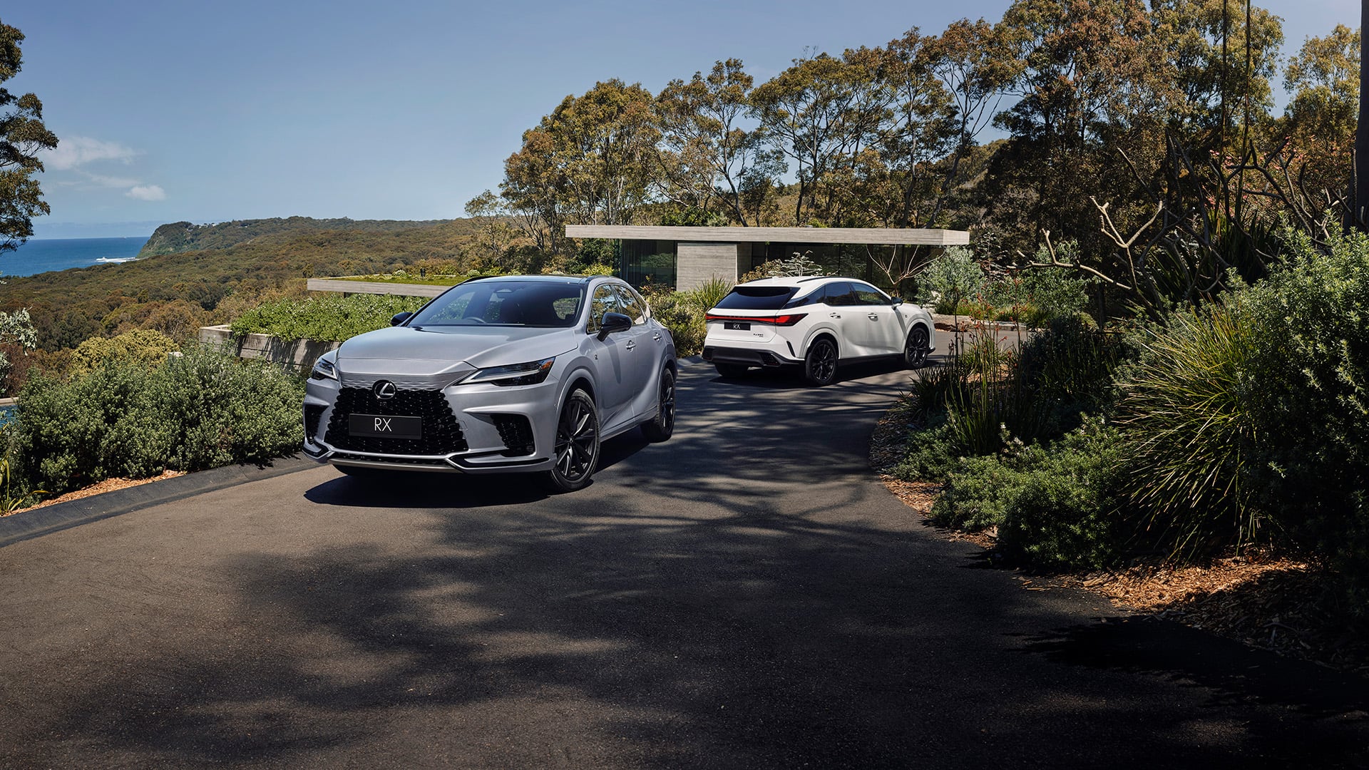 Two 2023 Lexus RX Vehicles are parked on a driveway in front of a luxury house. The sun is shining.